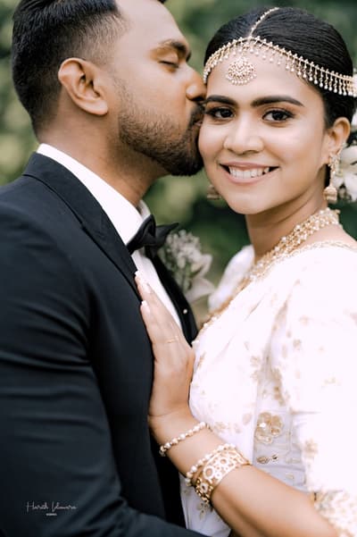 A groom kisses his bride on the forehead. She wears a white saree and a traditional headpiece.