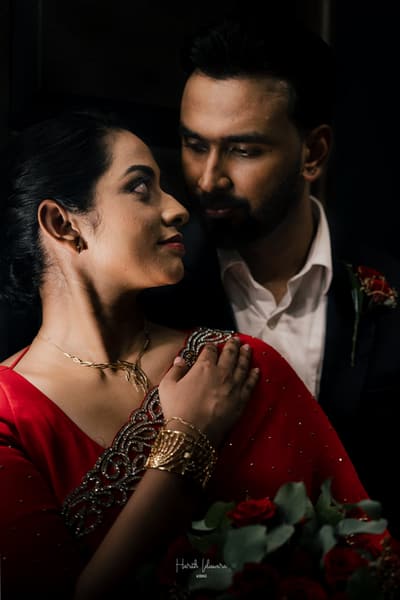 A close portrait in low light. The bride in a red saree, the groom just behind her.
